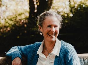 Pam Horton laughing and leaning against a gate, wearing white shirt and blue knitted cardigan