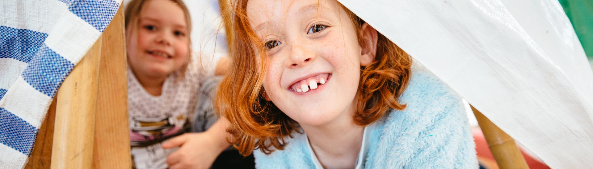 Two young children hiding in a hand made den and looking towards the camera