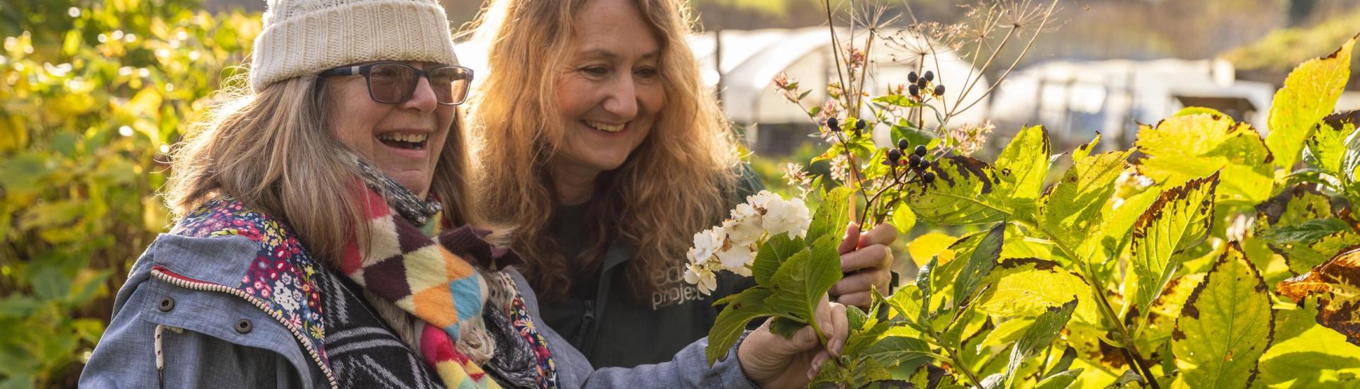 Two women, one wearing an Eden fleece, smile while looking at some plants outside. The weather is wintery, but there is a winter glow on the plants from the sun